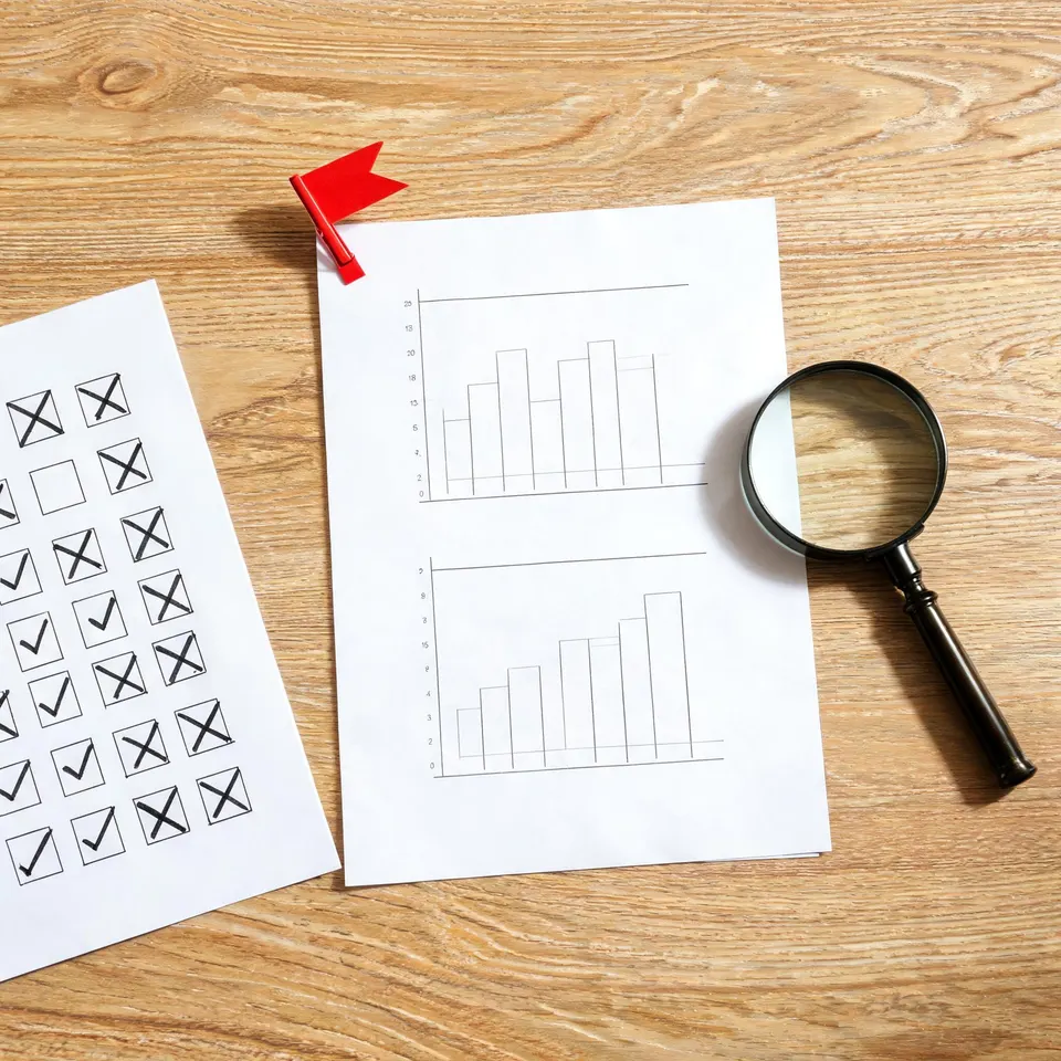 Flat-lay of audit report pages, red flag paperclip, magnifying glass and a crossed-out checklist on wood